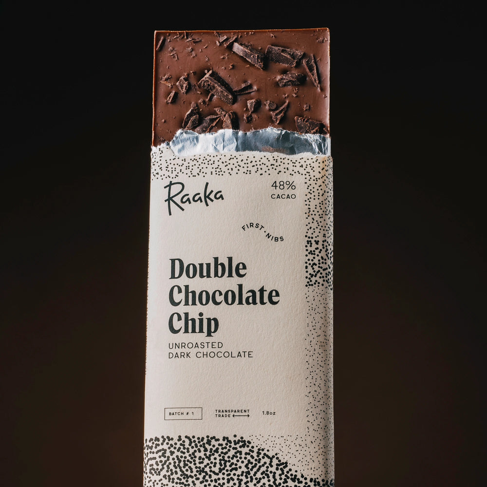 A single upright bar of Raaka Double Chocolate Chip in its paper wrapper, shown against a dark gradient background. The wrapper is cream-colored with black speckled patterns and text, and the top section of the chocolate bar is visible, showing a rich dark brown chocolate with chocolate shavings.