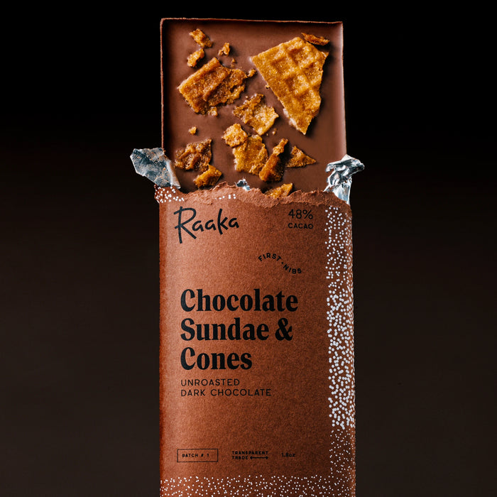 An open Raaka Chocolate Sundae & Cones bar in dark chocolate packaging, with broken waffle and cone-shaped chocolate pieces visible at the top, photographed against a dark background.
