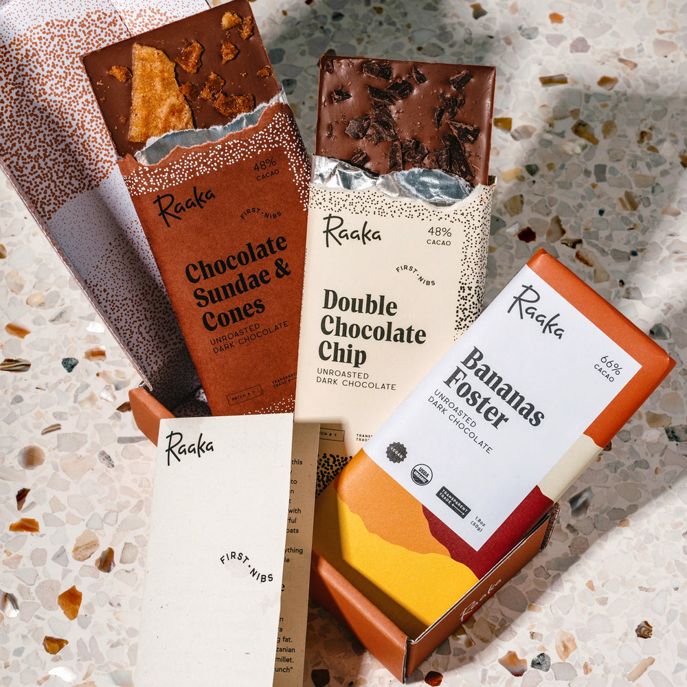 A set of RaaKa chocolate bars in their packaging, arranged on a speckled terrazzo surface. The visible bar types include Chocolate Sundae & Cones, Double Chocolate Chip, and Bananas Foster, with one bar partially unwrapped showing the chocolate texture.