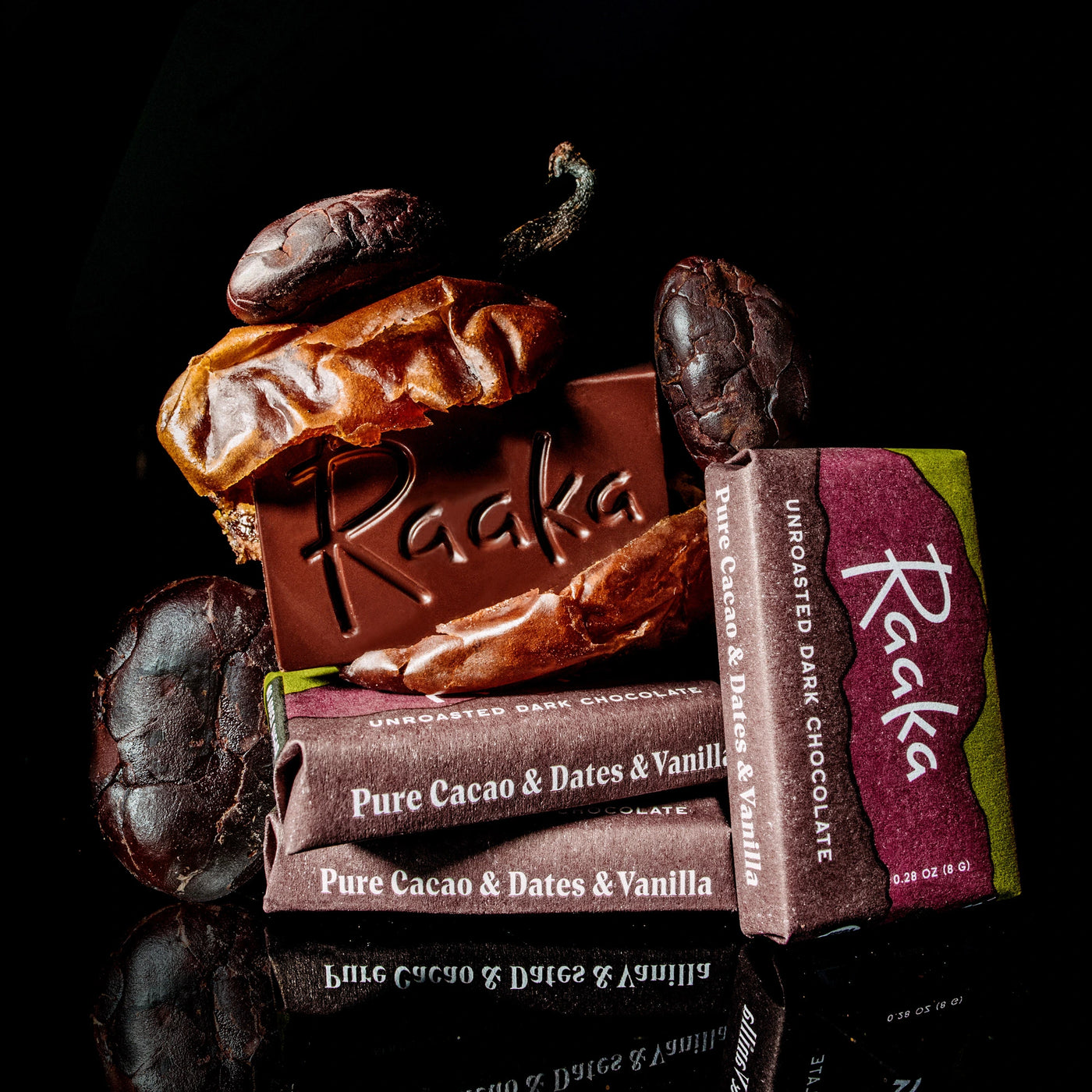 A mini chocolate bar embossed with the Raaka logo, two wrapped bars labeled Pure Cacao & Dates & Vanilla, and several dark cacao beans, a date, and vanilla bean.