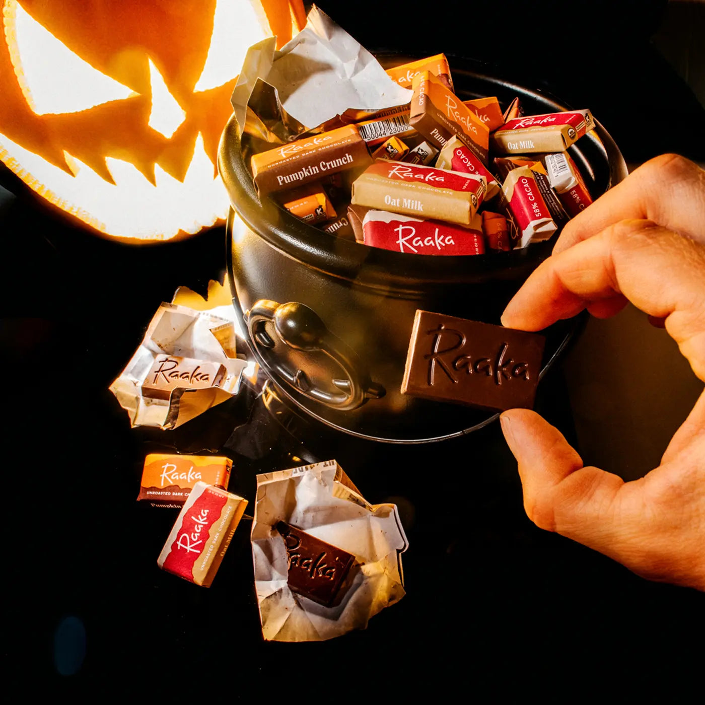 A cauldron of mini chocolate bars next to a jack-o-lantern. A hand is holding an unwrapped mini.