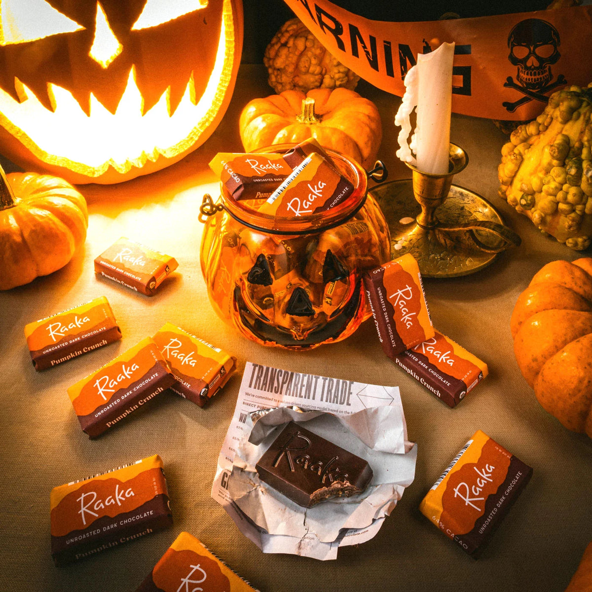  Halloween-themed photo of Raaka Pumpkin Crunch minis arranged around carved pumpkins, a candle, and seasonal props on a tabletop.