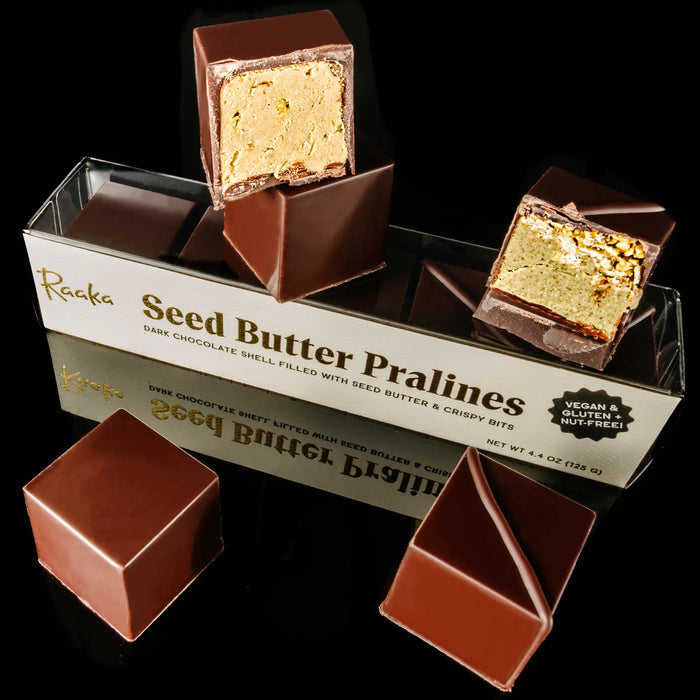 Six seed butter praline cubes stacked on a dark background with their packaging. Two are cut open to reveal a nougat-like center. The packaging has a label with metallic text.