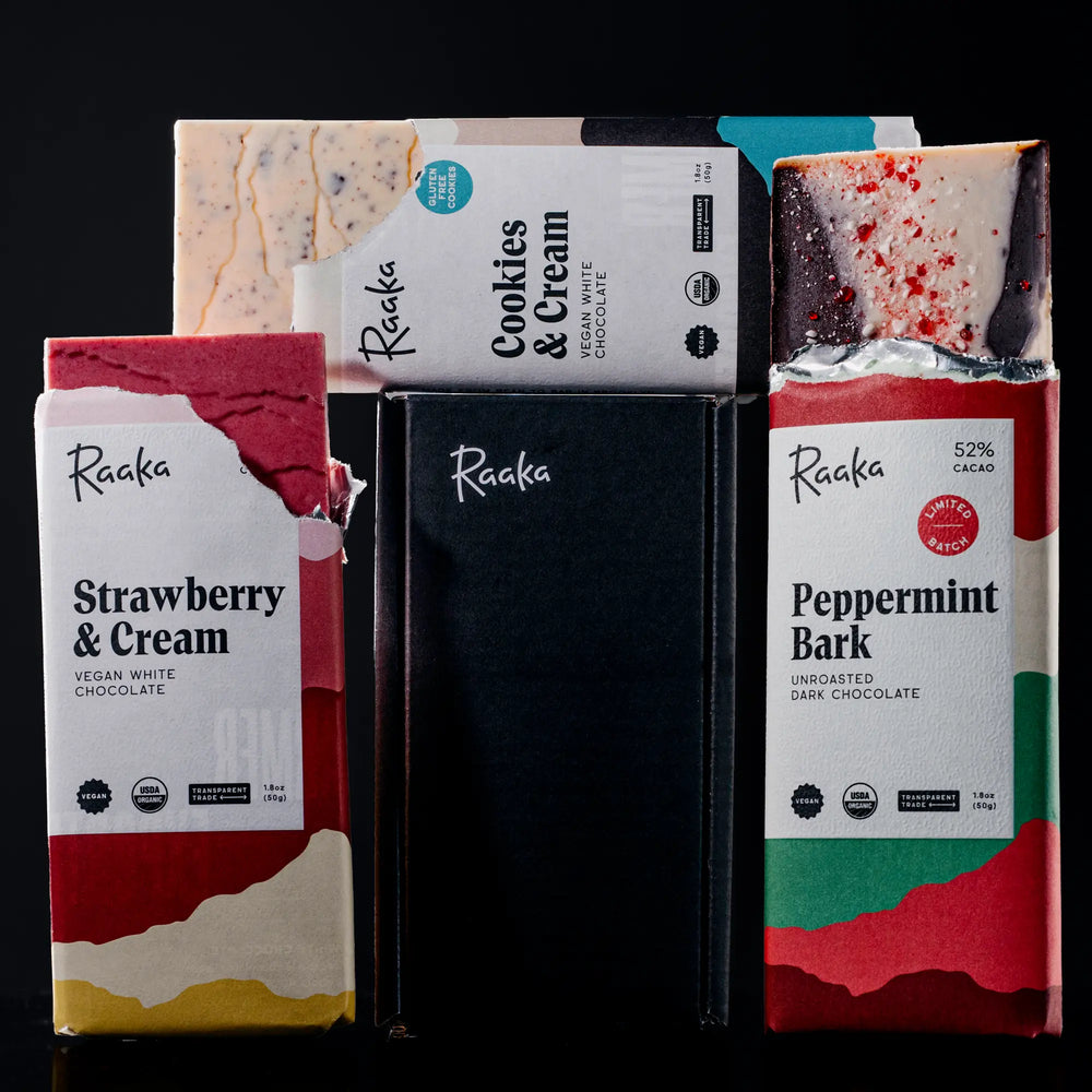 Three Raaka white chocolate bars on a black background, positioned around the box they come in: Peppermint Bark, Cookies and Cream, and Strawberry & Cream.