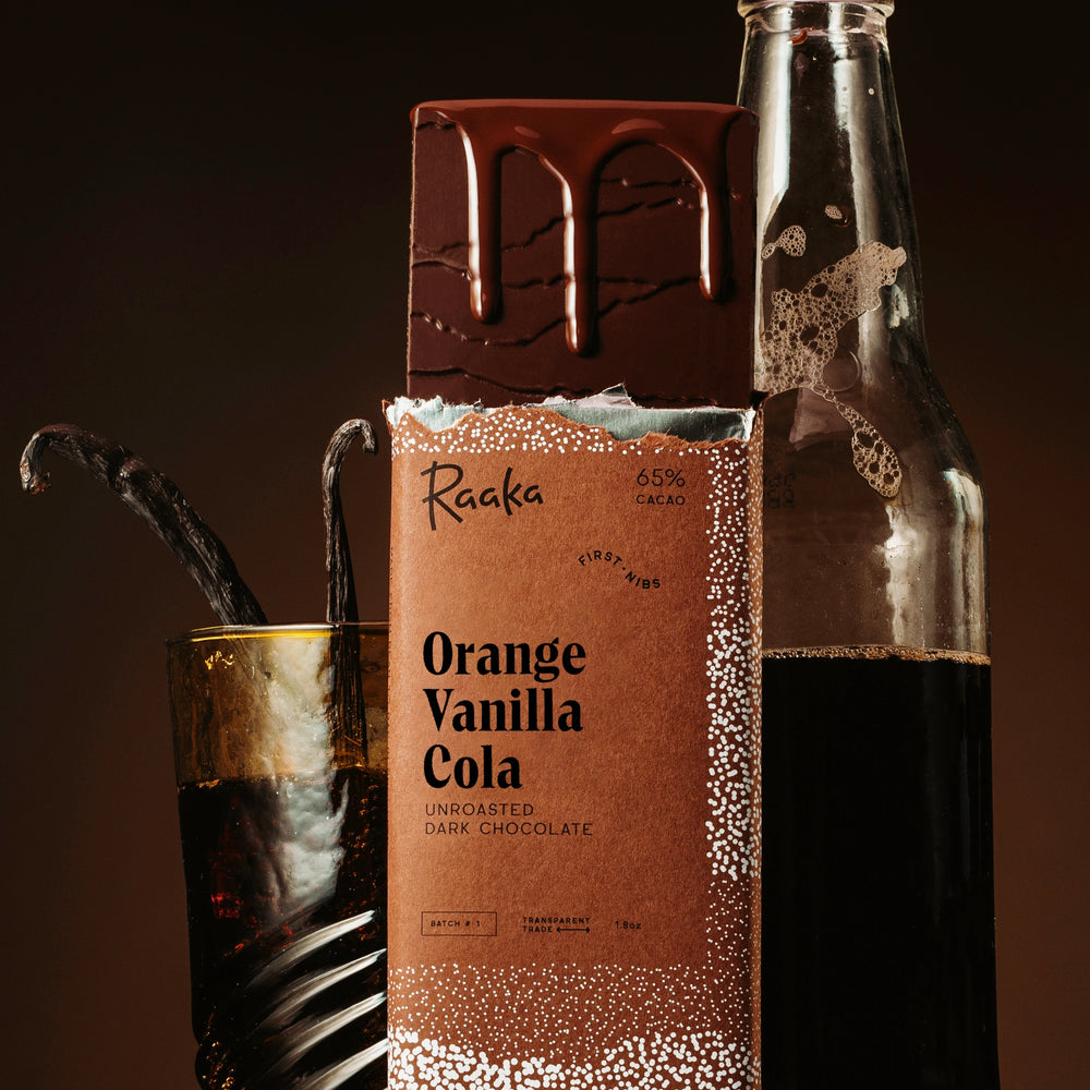 A bar of Raaka Orange Vanilla Cola dark chocolate stands upright in front of a glass bottle and a glass with vanilla beans, on a dark brown background.